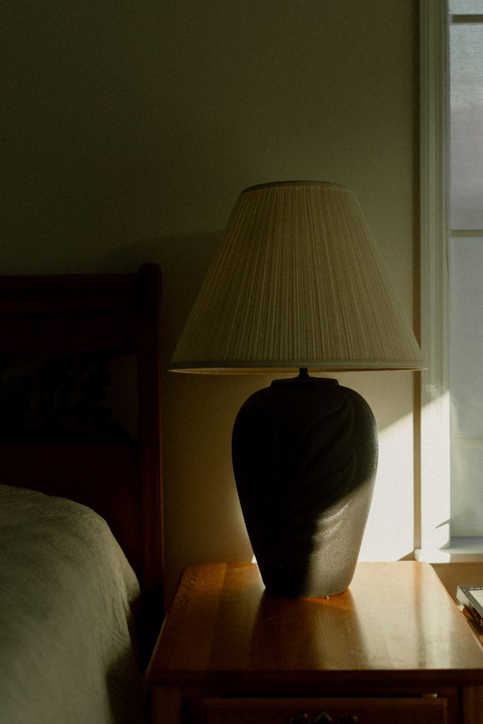 Warm sunlight illuminates a decorative lamp on a wooden nightstand in a cozy bedroom setting.